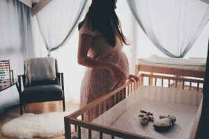 Pregnant woman in white lace sleeveless dress standing beside brown wooden crib photo